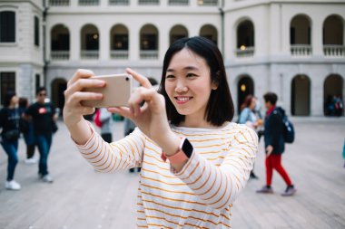 Neşeli etnik gezgin, gülümsüyor ve özçekim için poz veriyor. Şehir caddesindeki meydanın ortasında bulanık bir arka planda.