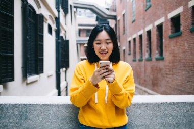 Şen şakrak kısa saçlı, sarı kapüşonlu bir kadın telefonda mesaj atıyor. Ekrana bakıyor, kentsel arka planda beton çite yaslanıyor. 