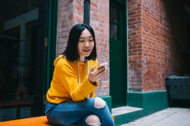 Genç, kısa saçlı, sarı kapüşonlu, yırtık pantolonlu, elinde telefonla ekranda oturan Asyalı kadın.