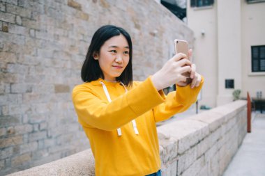 Neşeli, modern Asyalı kadın. Günlük kıyafetler içinde. Şehir sokağında tuğla duvarın önünde dururken akıllı telefondan selfie çekiyor.