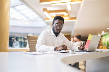 Görme düzeltmesi için göz giysisi giyen konsantre Afro-Amerikan erkek çalışanı. Finans raporlarını okuyor ve analiz ediyor. Ciddi, koyu tenli bir erkek yönetici, dizüstü bilgisayarla çalışma masasında oturuyor.