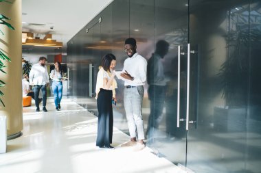 Çeşitli kadın ve erkek meslektaşlar birbirleriyle konuşuyor ve dijital tablette arkadaşça bir atmosferde kırılma videosu izliyorlar, olumlu koyu tenli erkek, gülümseyen kadın çalışana yeni bir cihaz sokuyor.