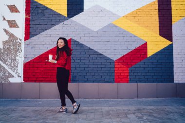 Renkli şehirlerde poz vermek için kahveyle gezen neşeli Asyalı genç kadın portresi, şık giyinmiş komik Japon milenyumu sokak sanatıyla turistik bölgeyi ziyaret ediyor.