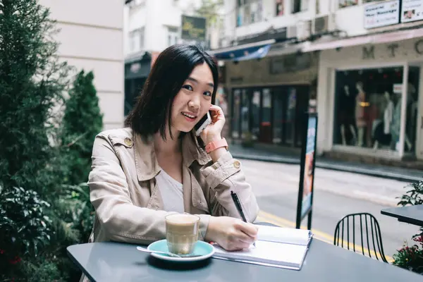 短い黒髪の若いアジアの女性は コーヒーのカップで机に座ってスマートフォンで話しながら 街道の背景にノートに書いた — ストック写真