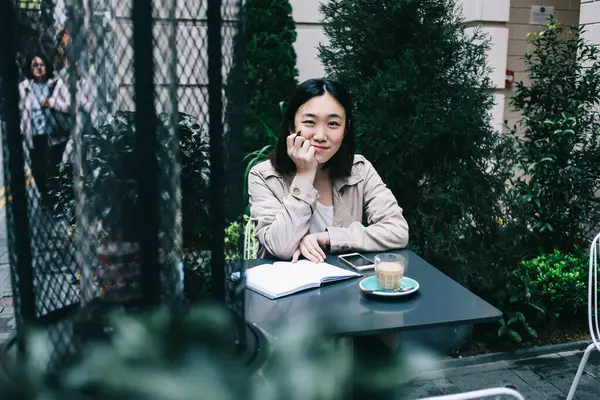 Joyful young Asian woman in casual clothes sitting at table with notebook and smiling while looking at camera and touching chin on street cafe near plants