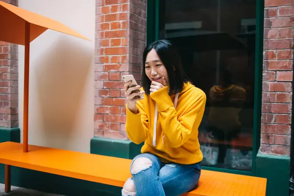 身穿黄色连帽衫的年轻亚洲女人坐在砖墙旁边的橙色长椅上 窗户很大 眼睛望着智能手机屏幕 — 图库照片