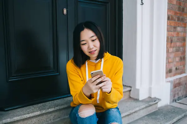身穿黄色帽衫的亚洲女人坐在门口的楼梯上 看着智能手机和打字留言 — 图库照片