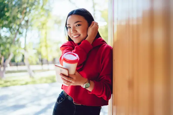 Half length portrait of pretty Asian millennial with smartphone and caffeine beverage standing at urban setting in city and smiling at camera, happy woman holding digital technology for browsing
