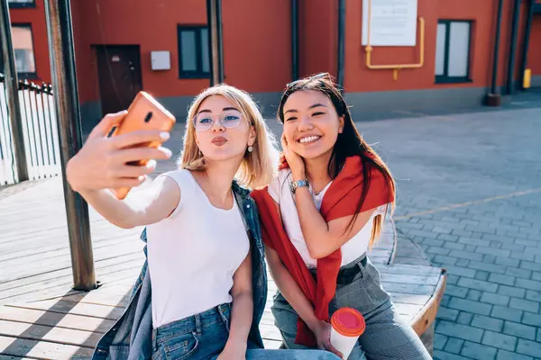 Joyful sincerely women having fun during friendly meeting at urban setting, cheerful millennials posing for making funny content images using front smartphone camera during leisure communication