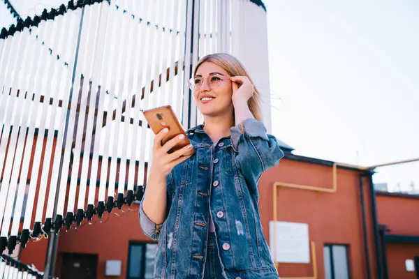 Cheerful hipster girl in optical eyewear for vision correction holding mobile standing at urbanity and smiling, carefree millennial woman blogger in trendy glasses using cellphone technology in city