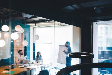 Koyu renk saçlı bir adamın cam penceresinden baktığında sakallı genç bir adama ve gözlüklü bir kadına fikir sunduğunu görüyoruz.