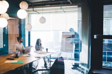 Beyaz tişörtlü, dizüstü bilgisayarlı, ofis malzemeli, modern iş yerinde koyu renk saçlı kadın sakallı bir adama fikir sunan işadamının penceresinden.