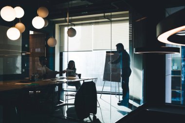 Büyük pencerelerin yanında duran ve elinde kağıt olan genç kadın ve erkeğe yeni bir fikir sunan bir iş adamı.