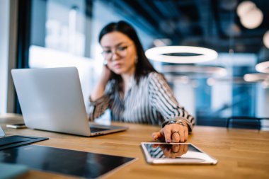 Koyu renk saçlı, gözlüklü, çizgili gömlekli bir kadın. Tabletin üzerindeki düğmeye basarken, modern iş yerindeki masada dizüstü bilgisayarla oturuyor.
