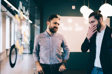 Beyaz gömlekli ve siyah ceketli sakallı adam, elleri cebinde koyu renk saçlı adamla gülüyor ve fikirleri tartışıyor.