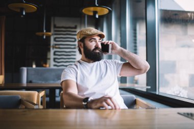 Modern barın ahşap masasında oturan şık şapkalı ve rahat gömlekli sakallı adam akıllı telefonla konuşuyor ve pencereden dışarı bakıyor.