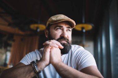 Ciddi sakallı, modaya uygun bir şapkası ve günlük kıyafetleri olan bir adam kafeteryada oturuyor. Elleri yüzüne yakın ve düşünceli bir şekilde başka tarafa bakıyor.