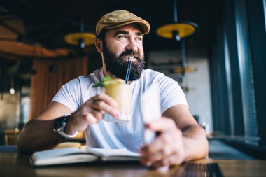 Sakallı, gülümseyen ve soğuk bir içecek içen erkek. Tatil sırasında defter ile çalışırken şık, modern, karanlık bir kafeye bakıyor.