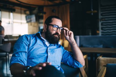 Ciddi sakallı bir adam, gözlüklü ve günlük kıyafetleriyle kafede oturuyor ve ara verip düşünürken dalgın dalgın dalgın bakıyor.