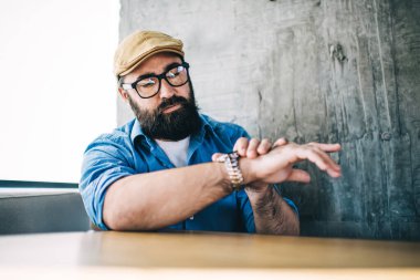 Gözlüklü, gözlüklü, sıradan kıyafetleri olan ve kafede otururken kol saatine bakan modaya uygun yetişkin bir hippi.