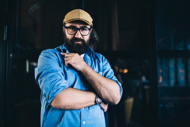 Kot gömlekli ve kepli hippi bir adam karanlık iç mekanda dikilirken kameraya ciddi ciddi bakıyor.