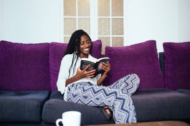 Gülümseyen koyu tenli, 20 'li yaşlarda bir kadın evde boş vakit geçirip ilginç hikayeler okuyor rahat koltukta oturuyor, Afro-Amerikalı güzel bir kadın evinde komik bir romana gülüyor.