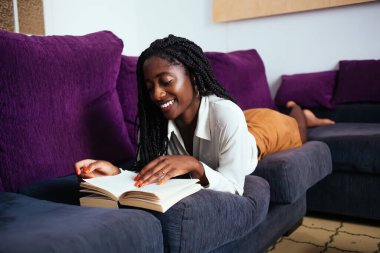 Neşeli koyu tenli kadın okuyucu oturma odasında kanepede uzanarak hobisine ve eğitimine vakit harcıyor. Afro-Amerikan 20 'li yaşlardaki yeniyetme kız rahat bir evde ilginç romanların sayfalarını çeviriyor.