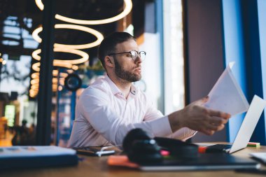 İnternet sitelerini taramak için modern dizüstü bilgisayarla masaüstü bilgisayarında çalışan finansal kağıt belgelerindeki görsel koruma araştırma bilgileri için optik gözlük takan akıllı serbest çalışan erkek.