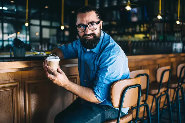 Male with beard in casual clothes sitting with cup of hot drink at wooden bar counter in trendy bar and looking away