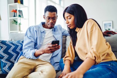 Afro-Amerikalı bir adam evde rahat bir oturma odasında kanepede otururken etnik kız arkadaşıyla akıllı telefondan bilgi paylaşıyor.