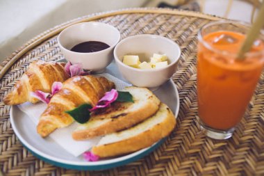 Lezzetli kruvasan tabağı ve reçel ve tereyağlı tost. Çiçek yapraklarıyla süslenmiş. Hasır masa üzerinde saman bulunan renkli bir içeceğin yanında.