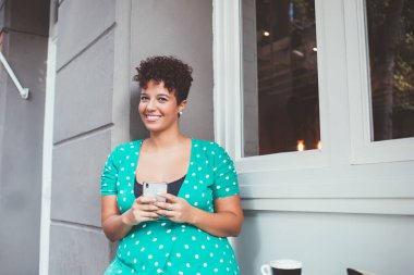 Aşırı neşeli Afro-Amerikalı kadın, günlük kıyafetli, gülümseyen ve kameraya bakan, açık kafedeki cep telefonundan bilgi arayan. 