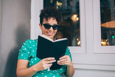 Neşeli gülümseyen, kıvırcık saçlı, günlük giysili, güneş gözlüklü, kafede dinlenen, neşeli, etnik bir bayan.