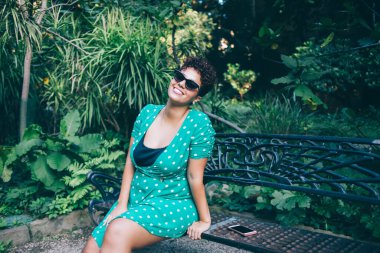 Güneş gözlüklü, sıradan elbiseli, siyah metal bankta tek başına oturan ve bahçede boş zamanlarında yeşil egzotik bitkilere karşı sırıtan güzel bir kadın.