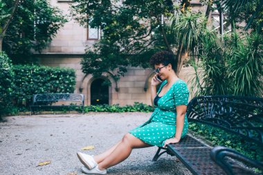 Güneş gözlüklü, yeşil noktalı yeşil elbiseli, siyah metal bankta otururken gün ışığında kemerli camları olan, bulanık eski binanın yanındaki yeşil egzotik bitkilere karşı telefon görüşmesi yapan düşünceli bir kadın.