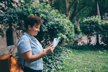 Sırt çantasıyla gezen, kağıt haritaya bakan ve parkta vakit geçiren hoş, etnik bir kadının yan görünüşü.