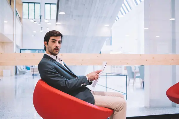 Portrait of South Asian male entrepreneur dressed in formal clothing looking at camera during time for analyzing documents, executive manager sitting in chair in modern workspace and posing