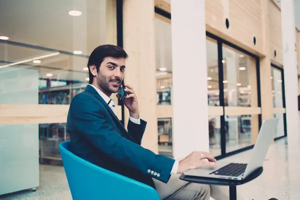 Portrait of successful South Asian employee in formal wear looking at camera during business smartphone conversation, confident corporate boss have cellphone consultancy call during laptop work