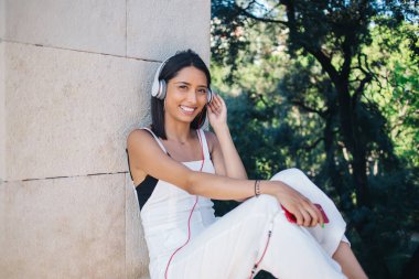 Modayı takip eden neşeli beyaz kadın portresi güneşli bir günde açık havada oturup müzik listesindeki müziklerle eğleniyor, kulaklıklarıyla gülümseyen bir kadın milenyumu şarkıları ve radyoları dinliyor.