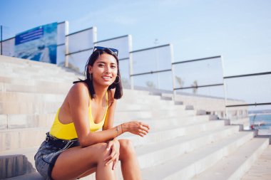 Parlak bluzlu ve şortlu neşeli etnik kadın şehir yürüyüşü sırasında dinlenirken taş basamaklarda oturuyor ve güneşin tadını çıkarıyor ve kameraya bakıyor.