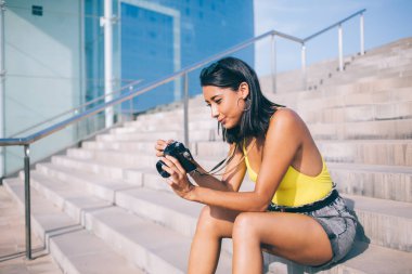 Tatil sırasında kentteki taş merdivenlerde otururken günlük kıyafet giymiş, fotoğraf makinesini yerleştiren tatmin olmuş etnik kadın gezginin yan görüntüsü
