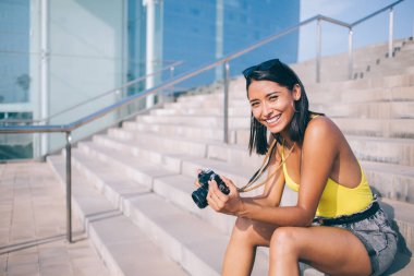 Neşeli bayan turist gülümseyerek ve fotoğraf makinesiyle taş basamaklarda otururken şehirde gezinirken ve kameraya bakarken.