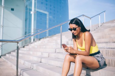 Haftasonu güneşli bir günde taş merdivenlerde dinlenirken güneş gözlüklü, modaya uygun giyinen ve cep telefonlu düşünceli bir etnik kadından.