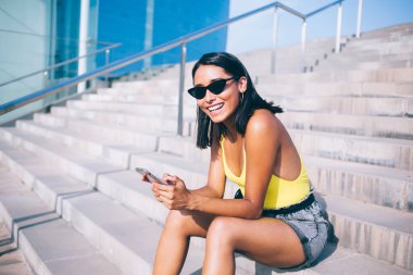 Yazlık kıyafetli, güneş gözlüklü, akıllı telefondan internette sörf yapan, şehirdeki taş merdivenlerde oturup kameraya bakan etnik bir kadın.