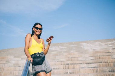 Günlük giysiler ve güneş gözlükleriyle sokakta taş merdivenlerde durup metal parmaklıklara yaslanırken akıllı telefondan sohbet eden etnik bir kadın.