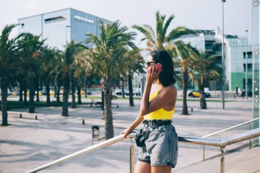 Kentte metal korkuluklara yaslanan ve merdivenlerde dikilip gözlerini kaçırarak cep telefonuyla konuşan şık etnik kadın.