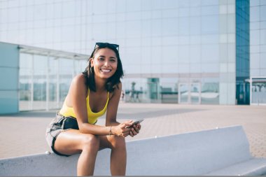 Yazlık kıyafetleri içinde, taştan bir bankta oturup kameraya bakarken sosyal medyada gülümseyen ve mesajlaşan pozitif etnik bir kadın.