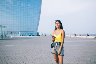 Modaya uygun giyinen, sosyal medyada cep telefonuyla sohbet eden ve şehrin modern cam binasının arka planında durup kameraya bakan etnik bir kadın.