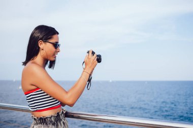 Moda kıyafetli düşünceli bir etnik kadın turistin deniz ufkunun arka planındaki setin üzerinde dikilirken fotoğraf çekerken çekilmiş fotoğraflarının yan görüntüsü.