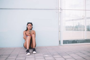 Cam duvara yaslanmış, taş zeminde oturmuş akıllı telefonlara bakarken ve kulaklıkla müzik dinlerken etnik kadın milenyumu.